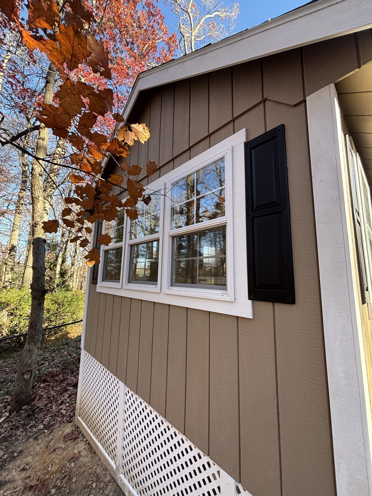 Exterior corner of a finished addition with brown siding, white-trimmed windows, and black shutters against blue sky. Home addition project by Potts Brothers Construction, Arlington & Northern Virginia