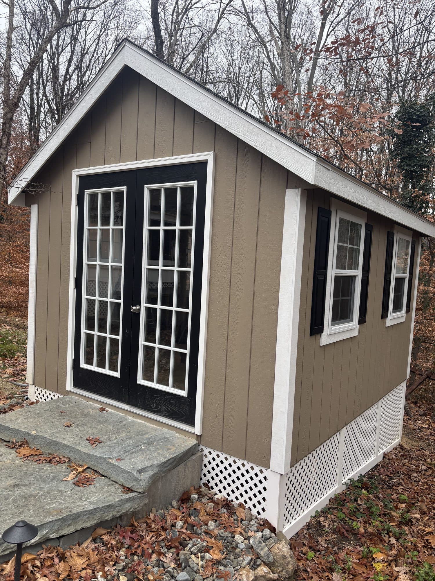 A finished taupe shed with white French doors, black shutters, and a lattice skirt beside a stone step. Home addition project by Potts Brothers Construction, Arlington & Northern Virginia