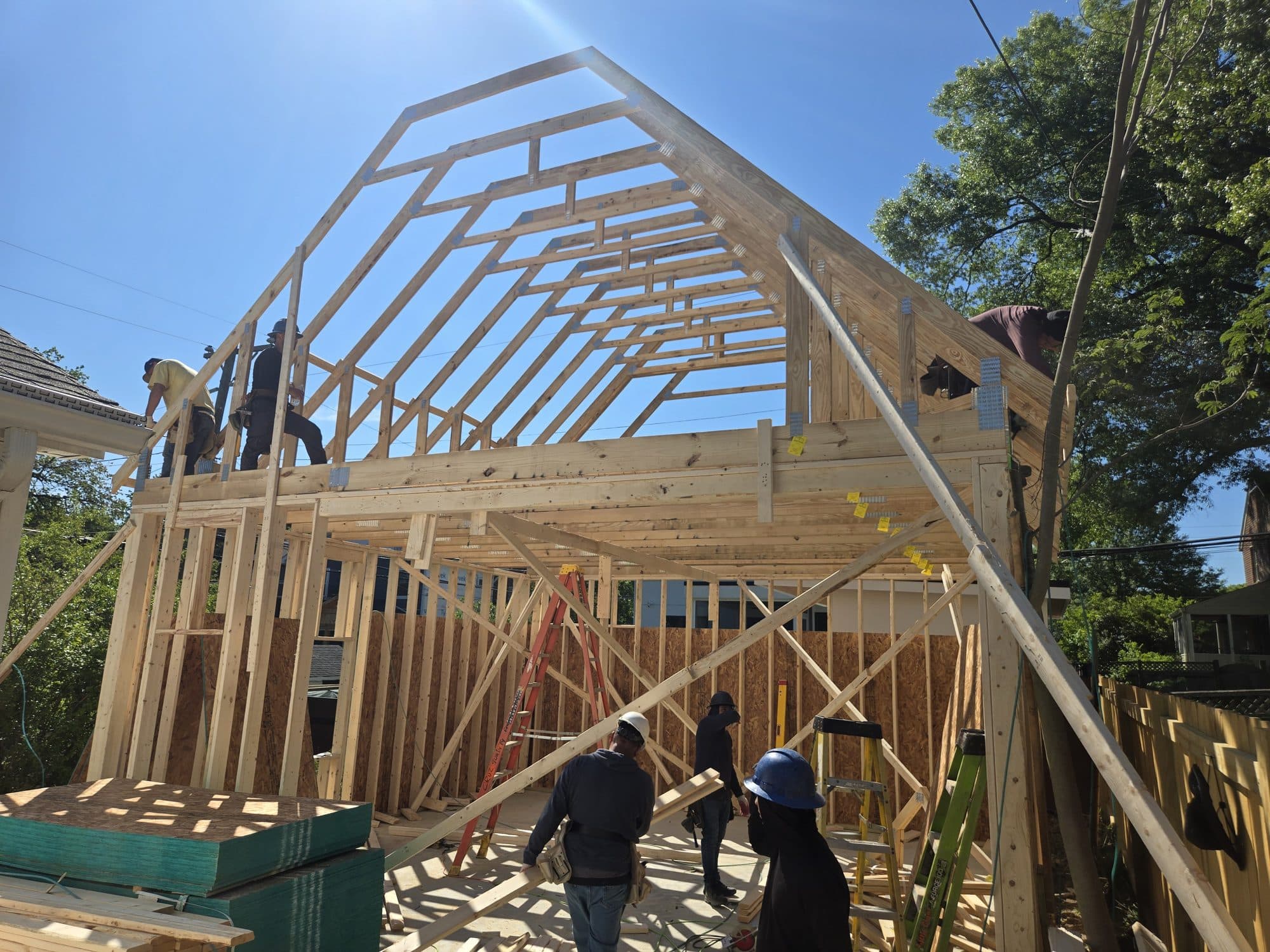 A crew raises gambrel rafter trusses over a newly framed two-story addition under a clear sky. Home addition project by Potts Brothers Construction, Arlington & Northern Virginia