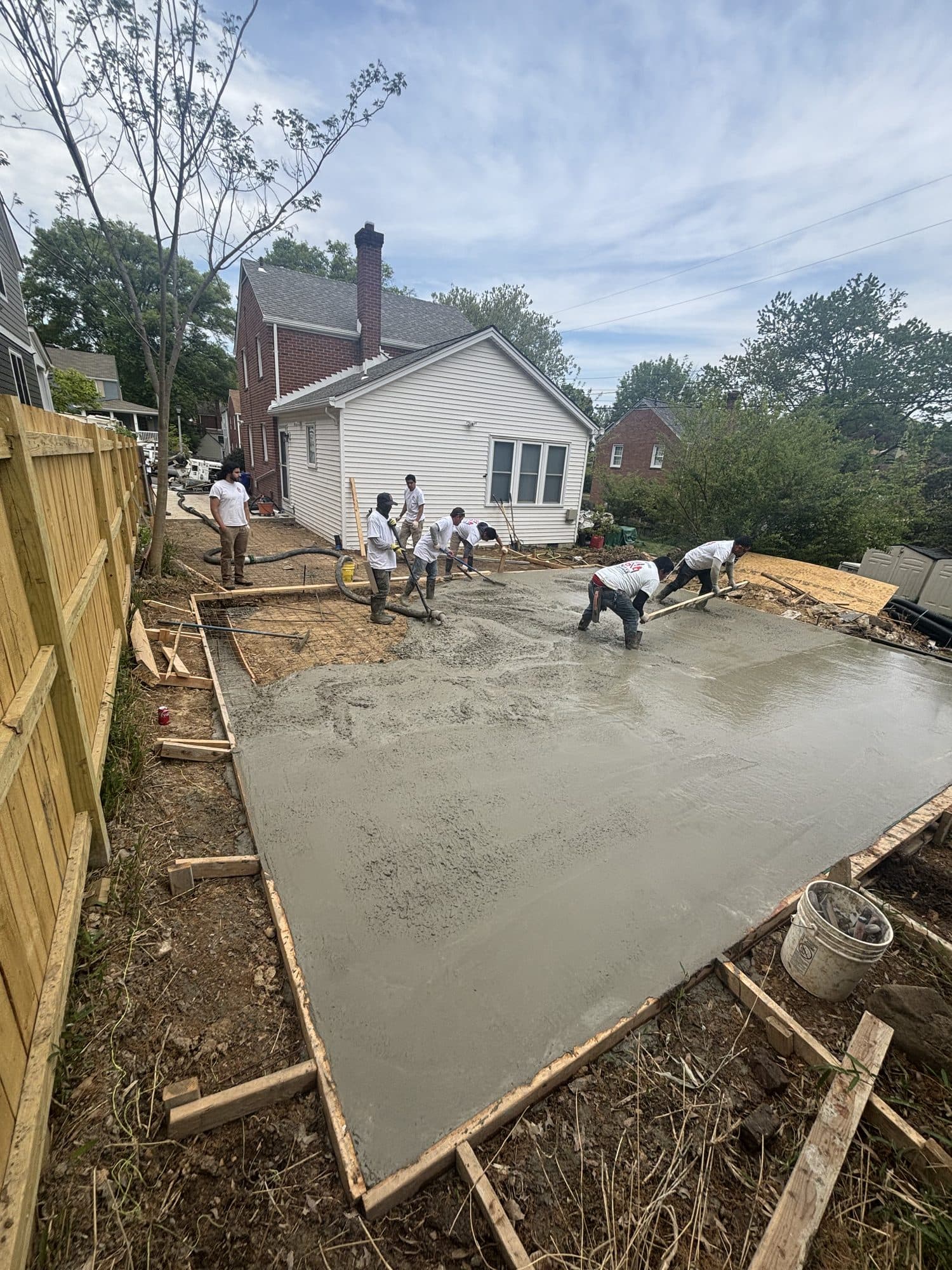 Workers screed and finish a large freshly poured concrete slab in a fenced backyard beside a white house. Concrete project by Potts Brothers Construction, Arlington & Northern Virginia