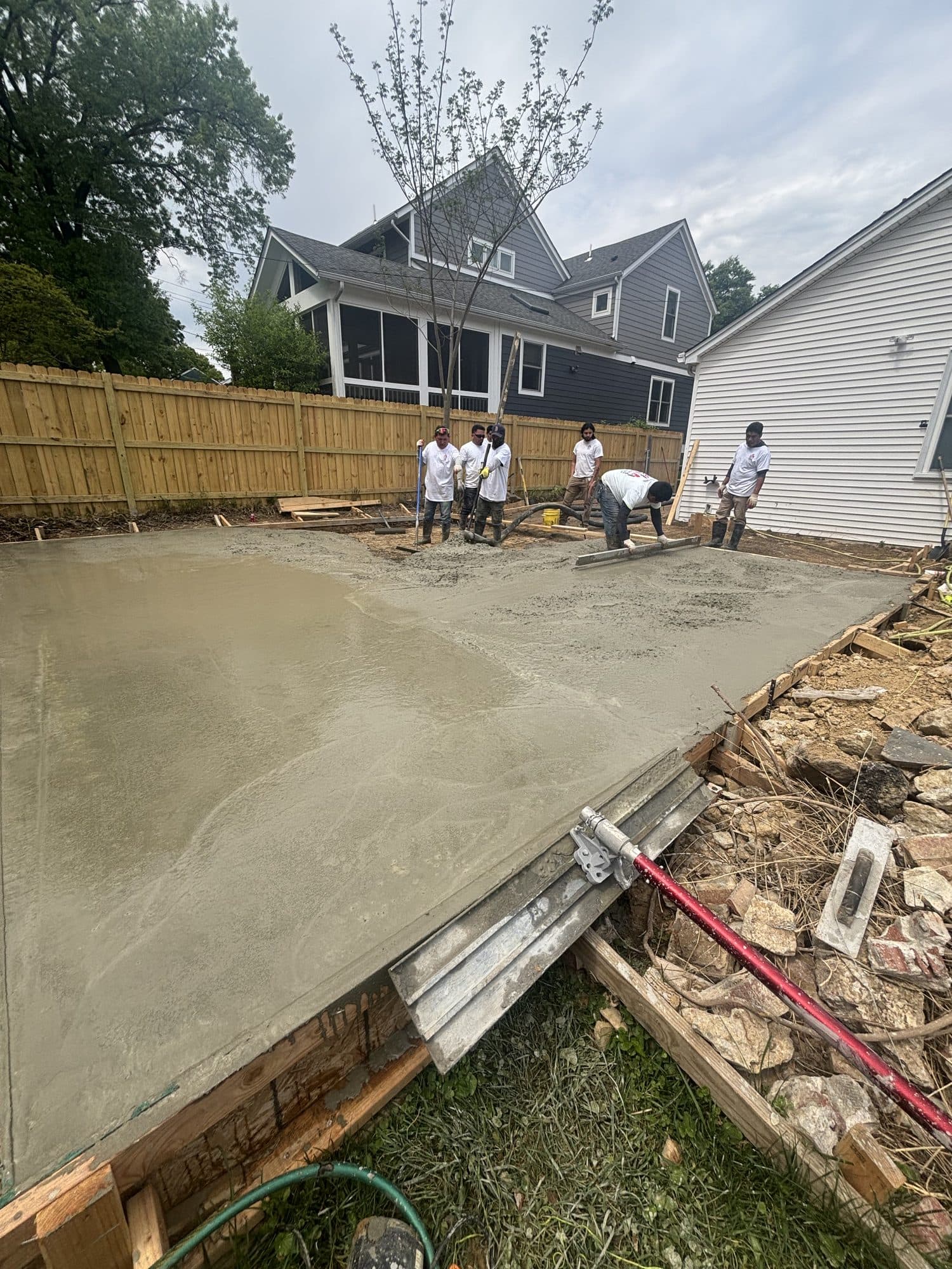 Workers float and finish a freshly poured backyard concrete slab edged by wood forms behind residential homes. Concrete project by Potts Brothers Construction, Arlington & Northern Virginia