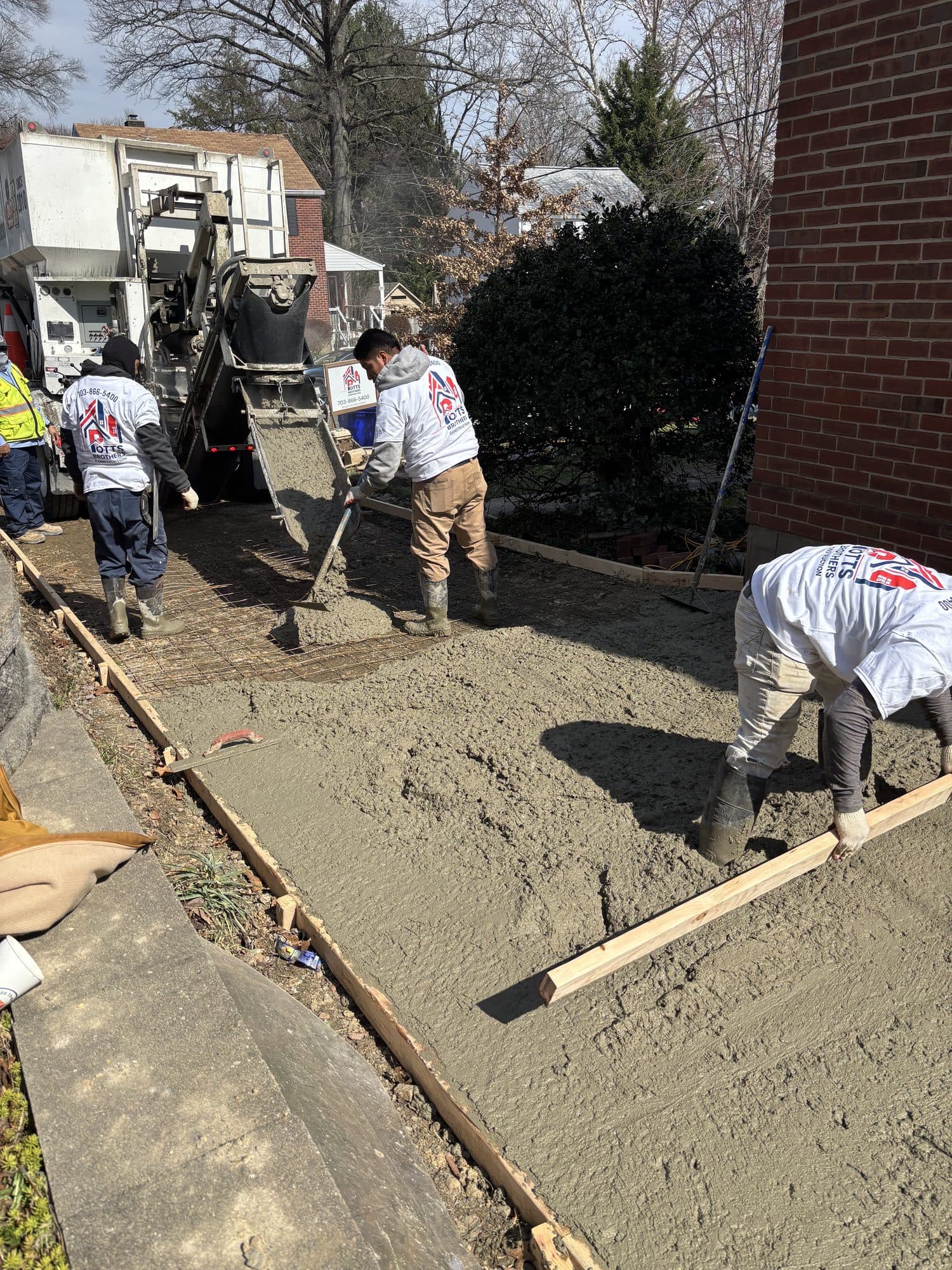 Workers spread and float wet concrete within a wood-formed slab beside a brick home with a concrete pump. Concrete project by Potts Brothers Construction, Arlington & Northern Virginia