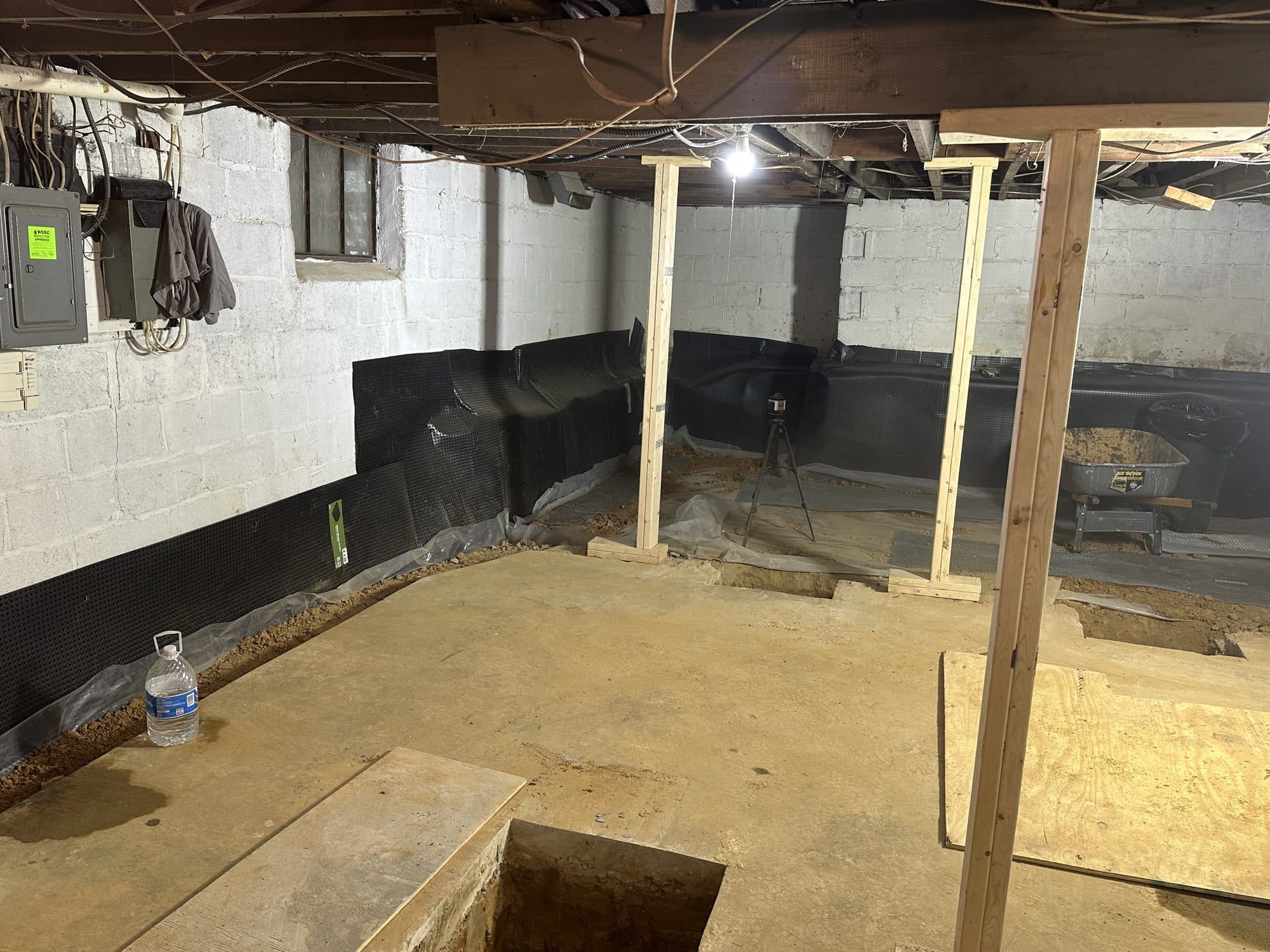 Basement interior showing a freshly poured concrete floor, temporary wood posts, dimpled drainage membrane along block walls, and an electrical panel. Foundation project by Potts Brothers Construction, Arlington & Northern Virginia