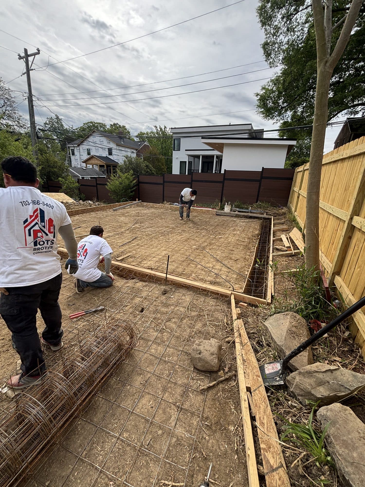Workers set wire mesh reinforcement inside wood forms preparing a backyard concrete slab beside a modern home. Foundation project by Potts Brothers Construction, Arlington & Northern Virginia