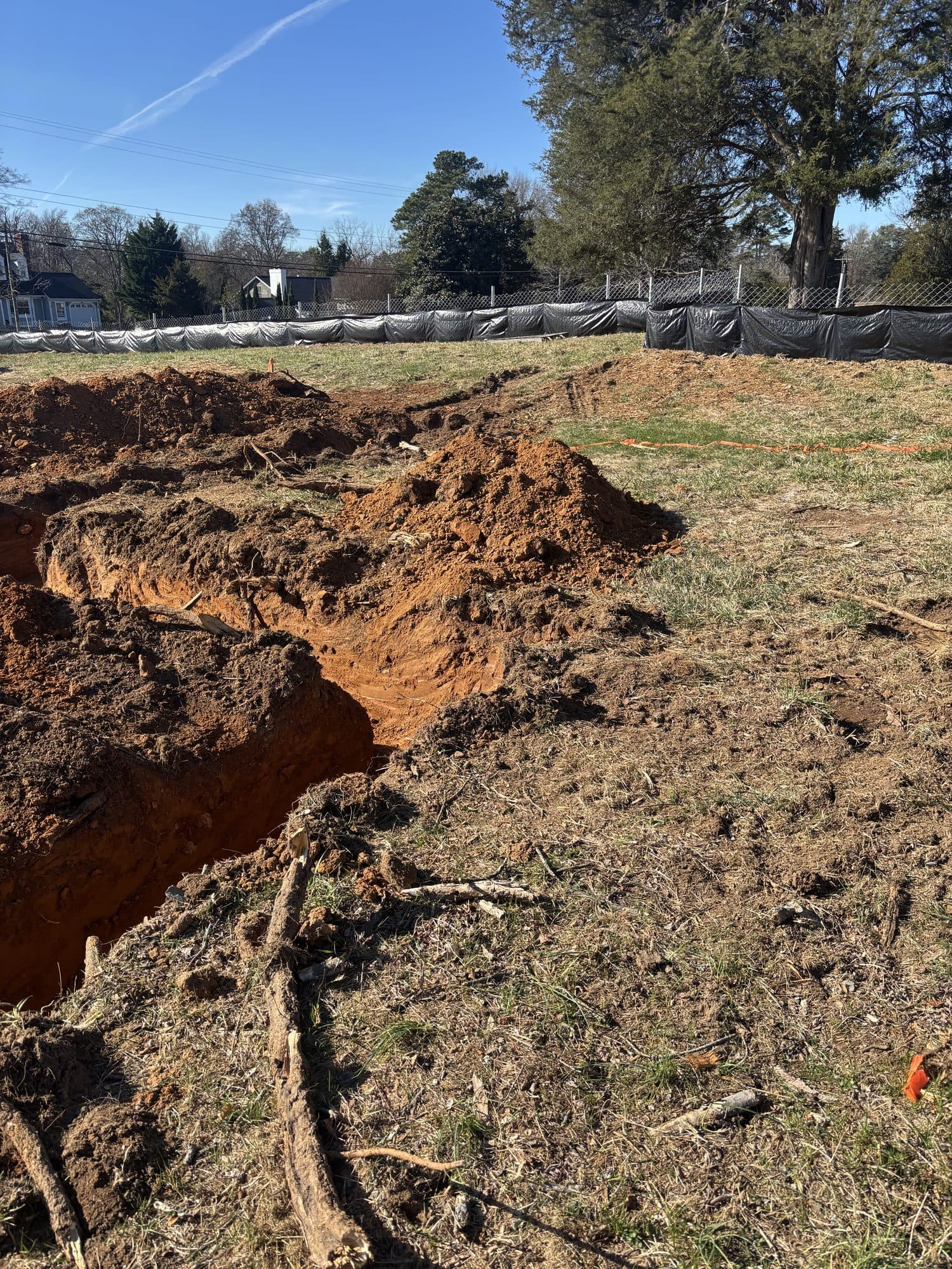 A freshly dug foundation trench cut through red clay soil on a fenced residential lot. Foundation project by Potts Brothers Construction, Arlington & Northern Virginia