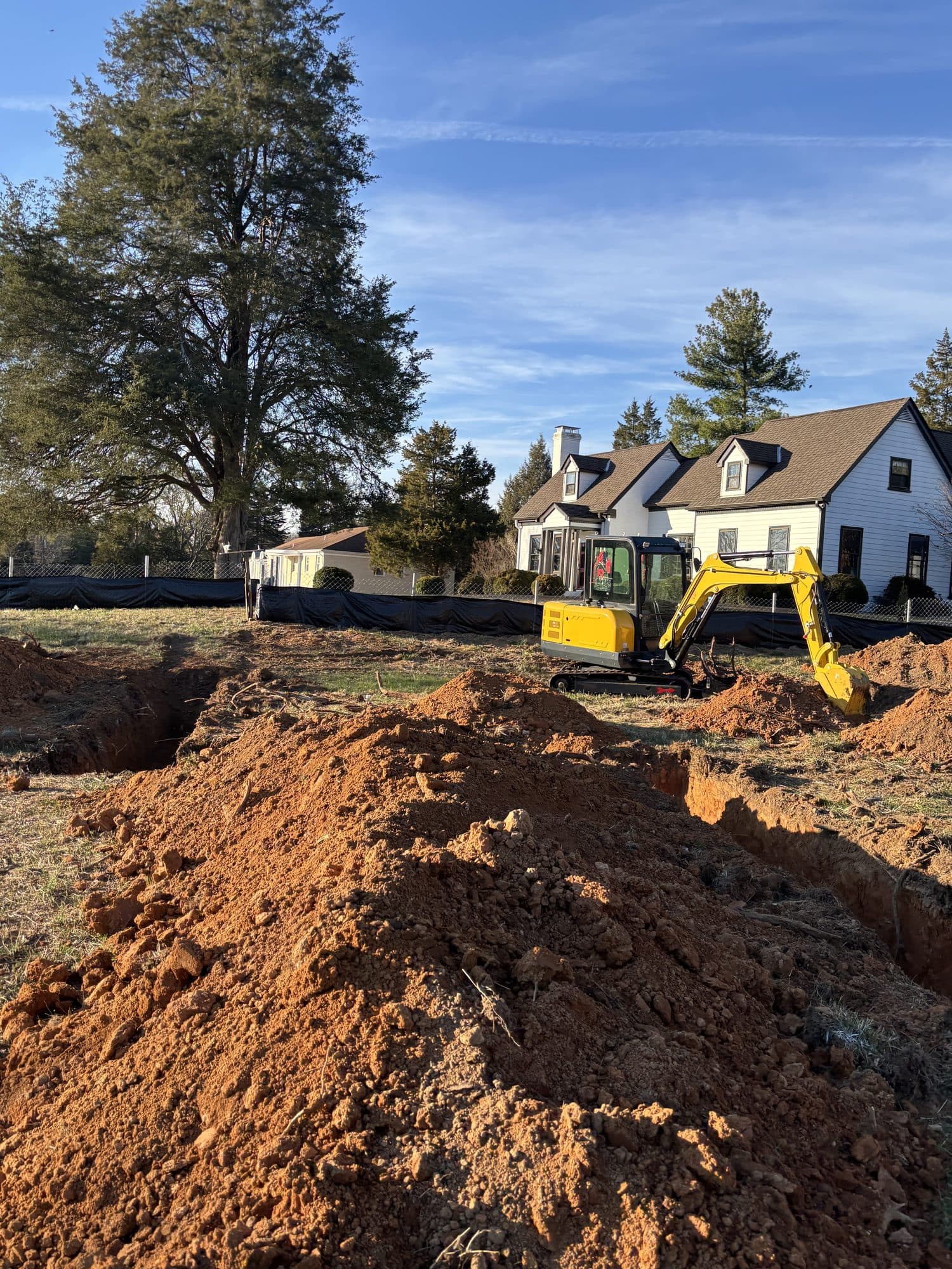 A yellow excavator beside a dug foundation trench in red clay soil with a white house behind. Foundation project by Potts Brothers Construction, Arlington & Northern Virginia
