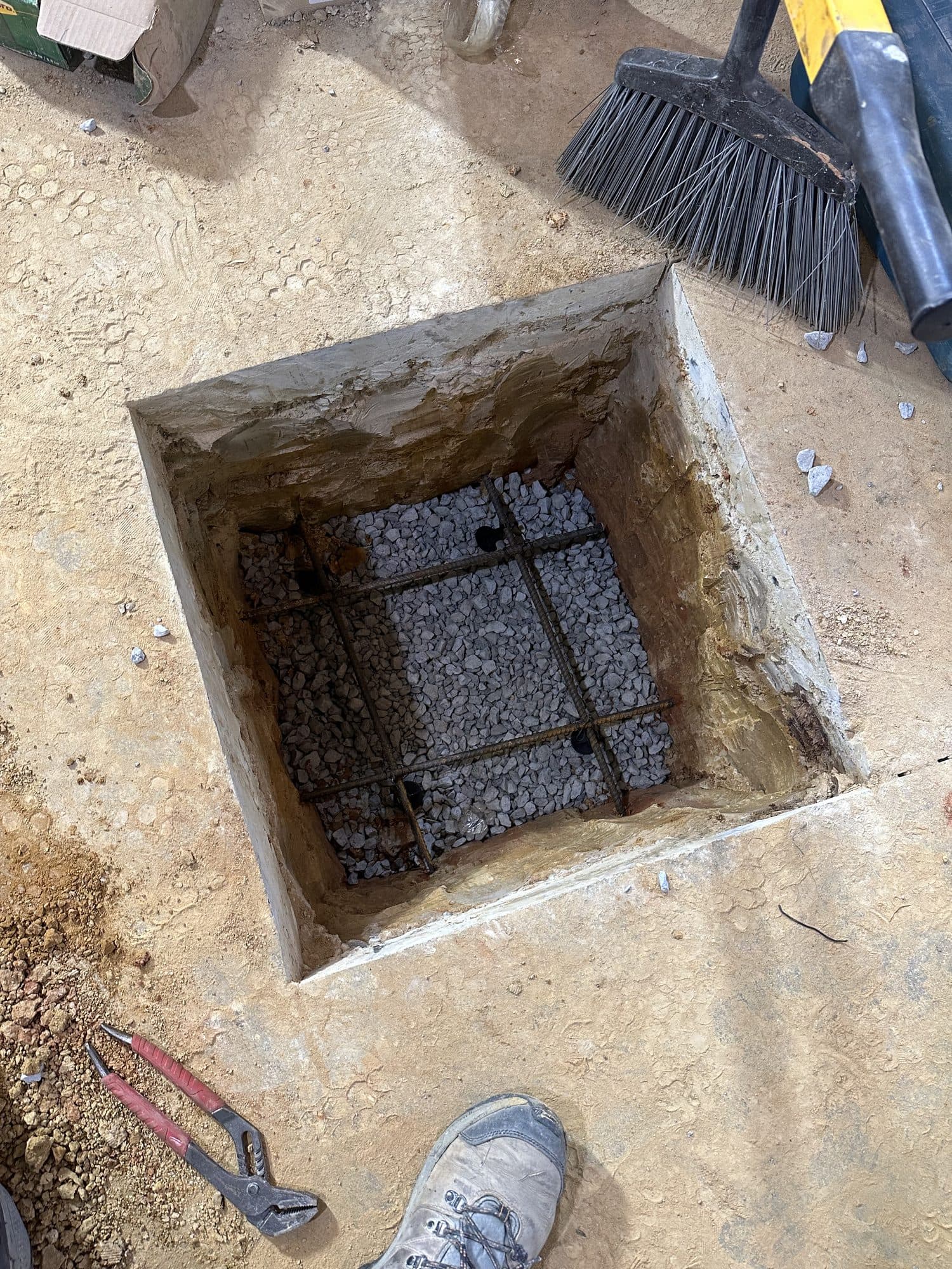 A square footing pit cut into a concrete slab containing a tied rebar cage over gravel, ready for a poured concrete pier. Foundation project by Potts Brothers Construction, Arlington & Northern Virginia