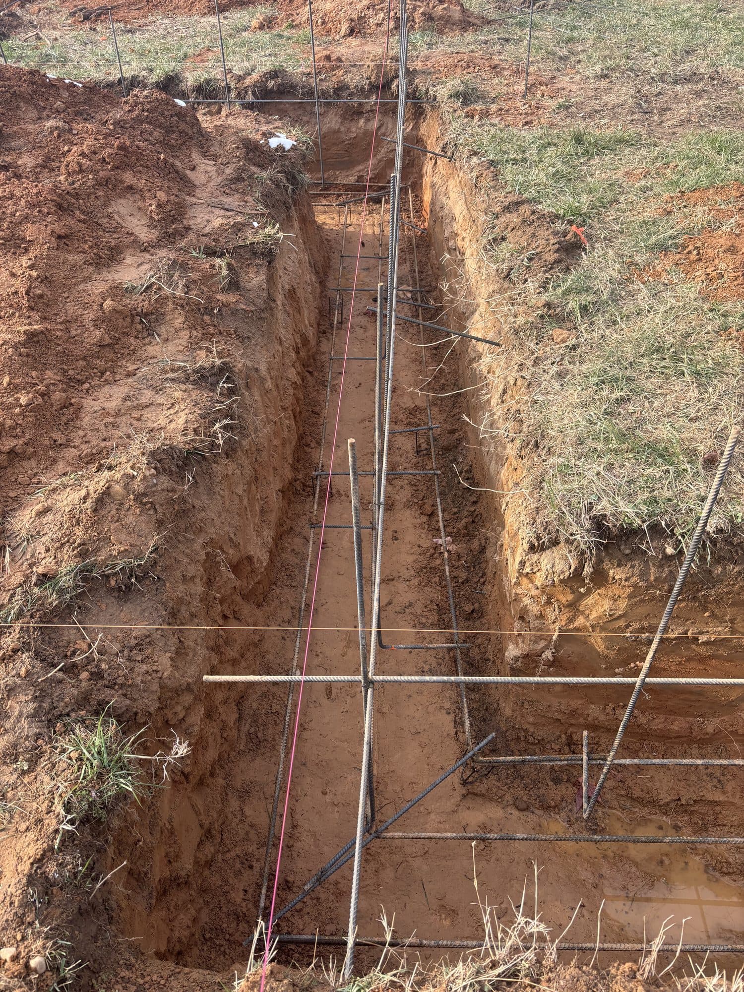 A foundation footing trench with rebar and a company yard sign beside a compact track loader. Foundation project by Potts Brothers Construction, Arlington & Northern Virginia