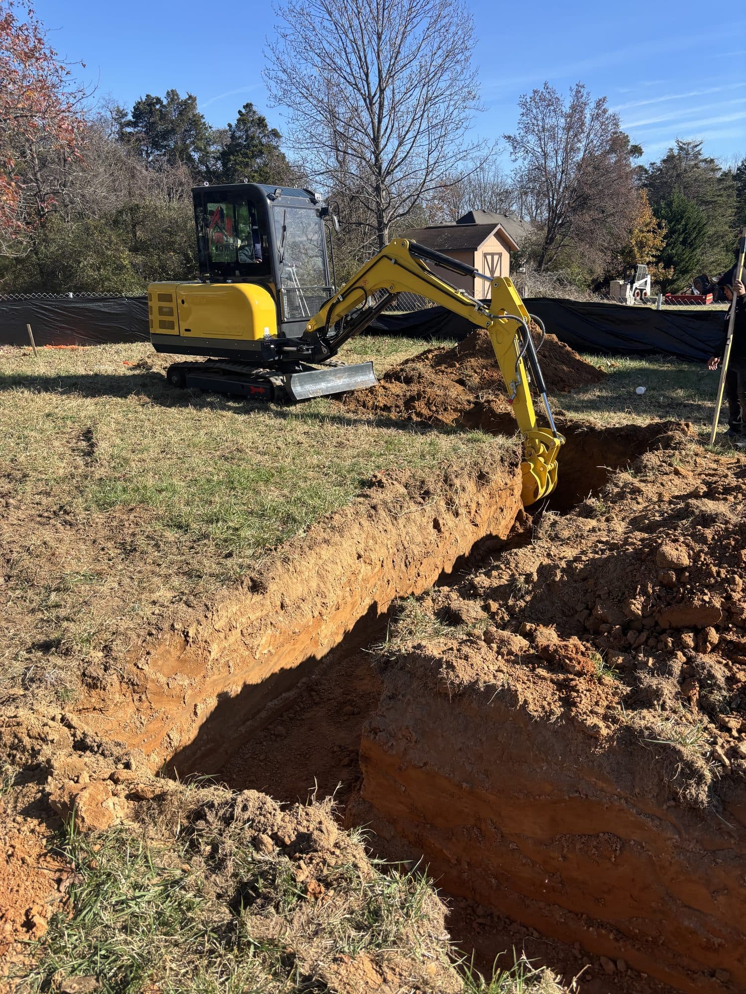 A yellow mini excavator digging a deep footing trench in a grassy yard with a shed behind. Foundation project by Potts Brothers Construction, Arlington & Northern Virginia