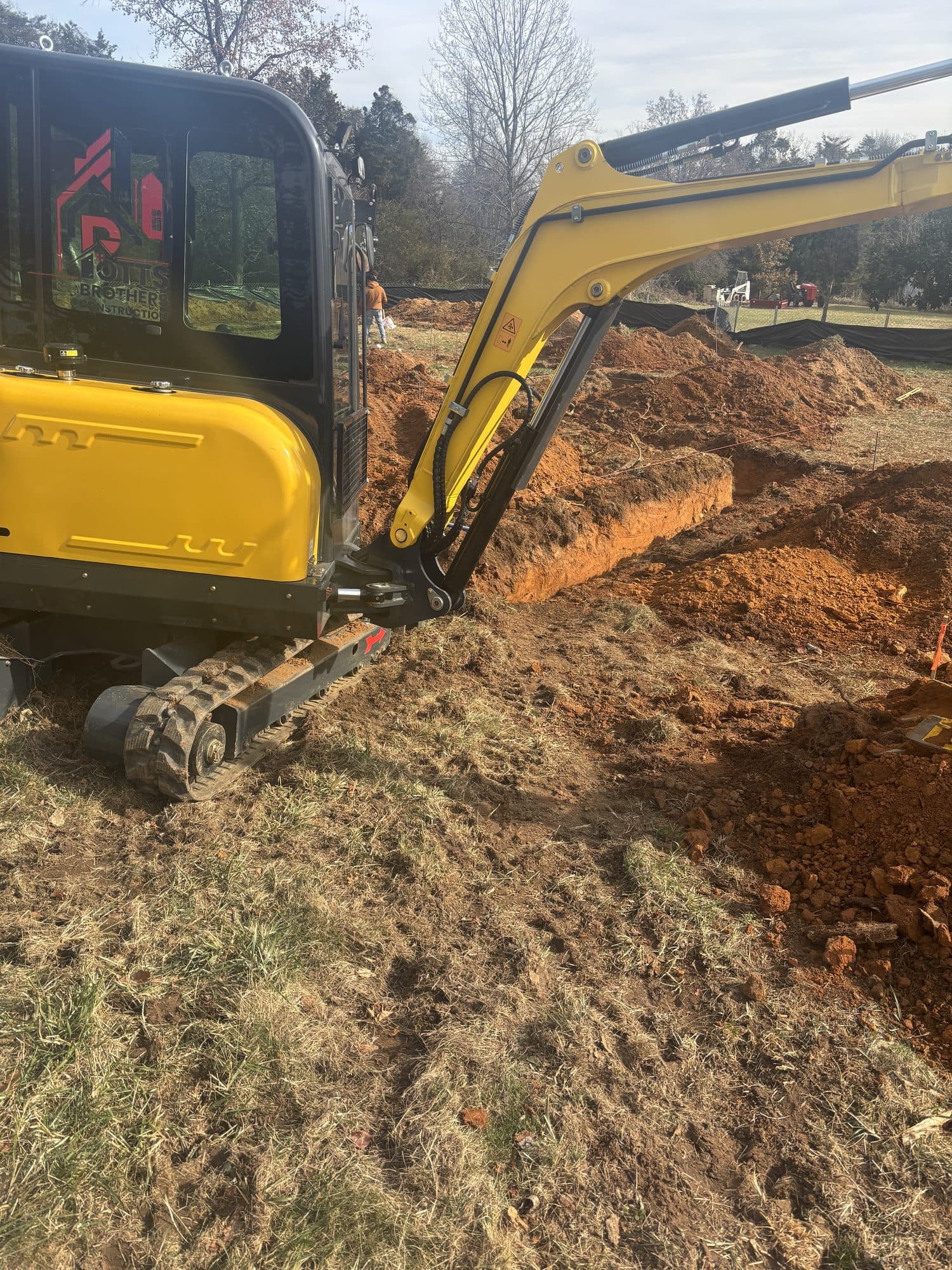 A yellow tracked mini excavator digging along the edge of a foundation trench in a yard. Foundation project by Potts Brothers Construction, Arlington & Northern Virginia