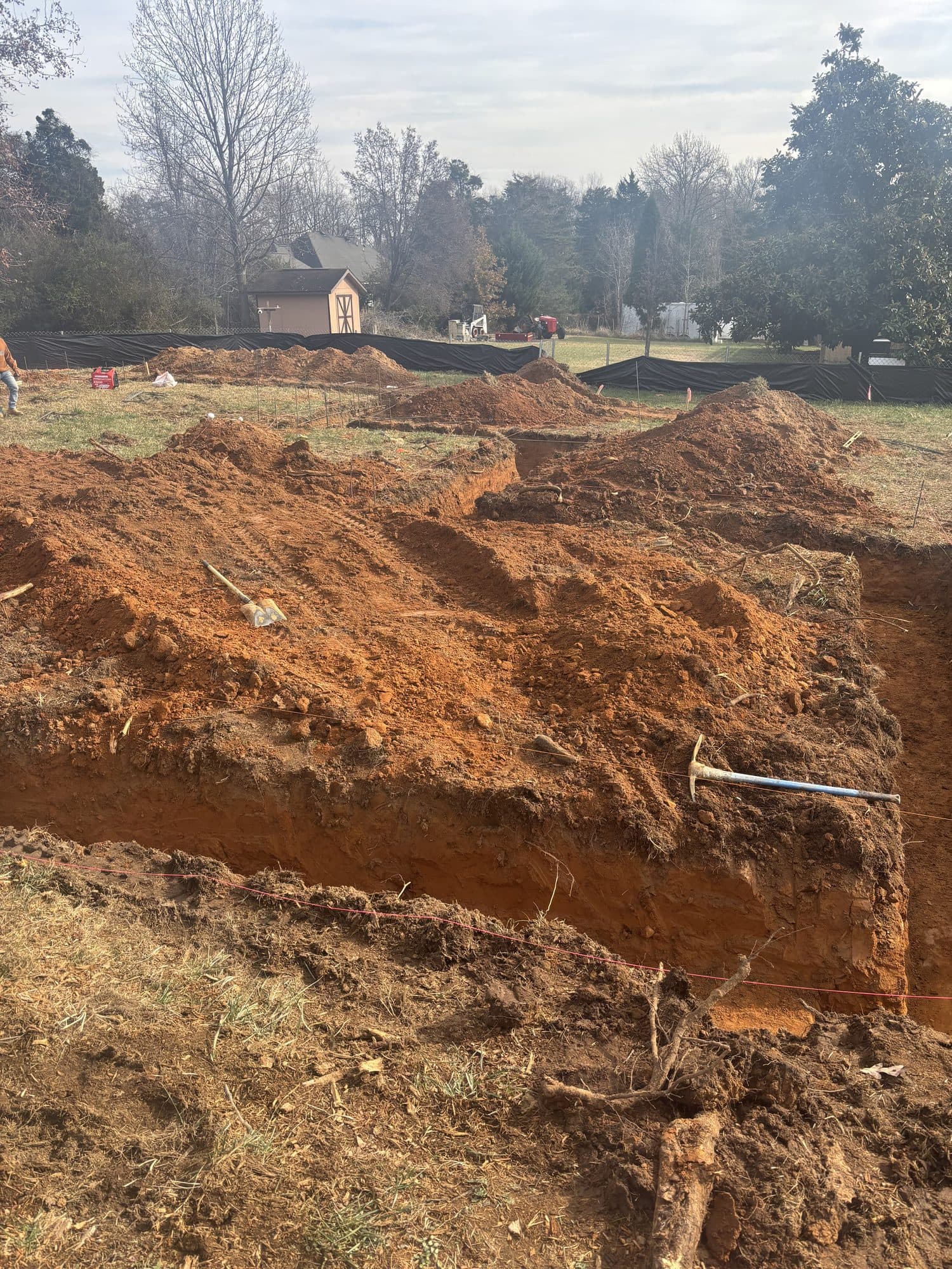 A wide foundation excavation with stepped footing trenches cut into red clay on a residential lot. Foundation project by Potts Brothers Construction, Arlington & Northern Virginia