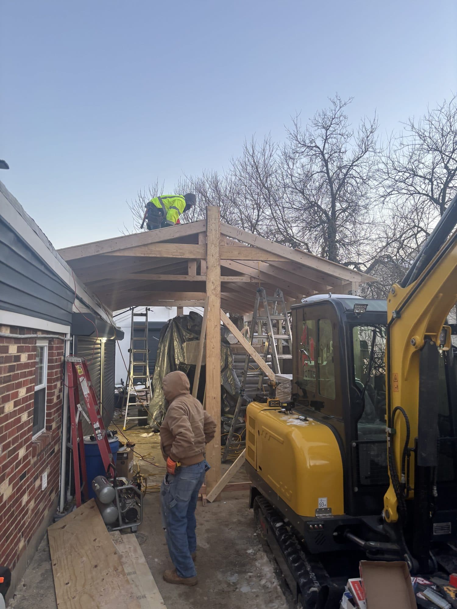 A crew frames the rafters of a detached garage while a compact excavator works alongside the open timber structure. Garage project by Potts Brothers Construction, Arlington & Northern Virginia