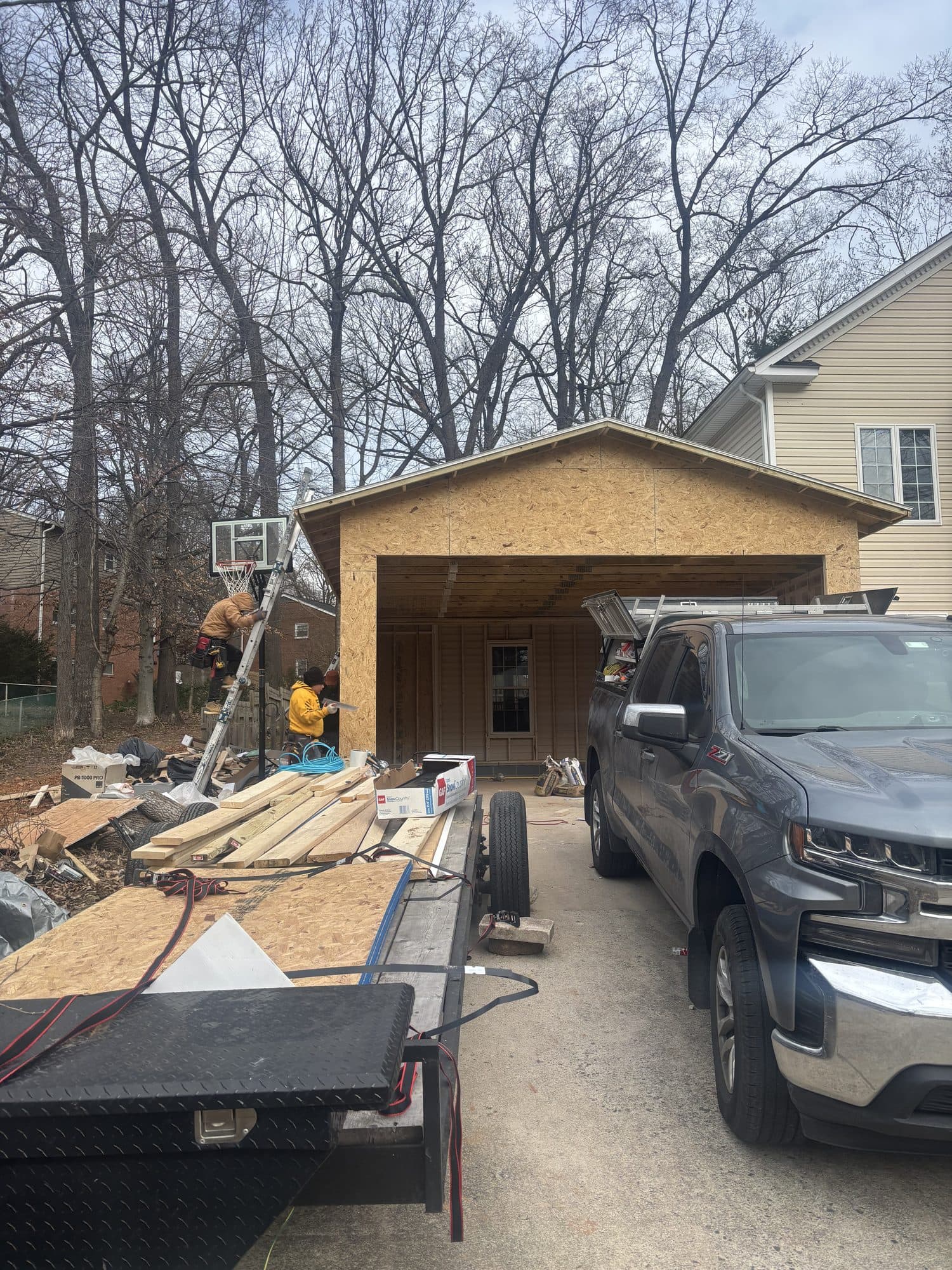 A detached garage with an open bay freshly sheathed in OSB next to a finished home, framing lumber staged in the driveway. Garage project by Potts Brothers Construction, Arlington & Northern Virginia