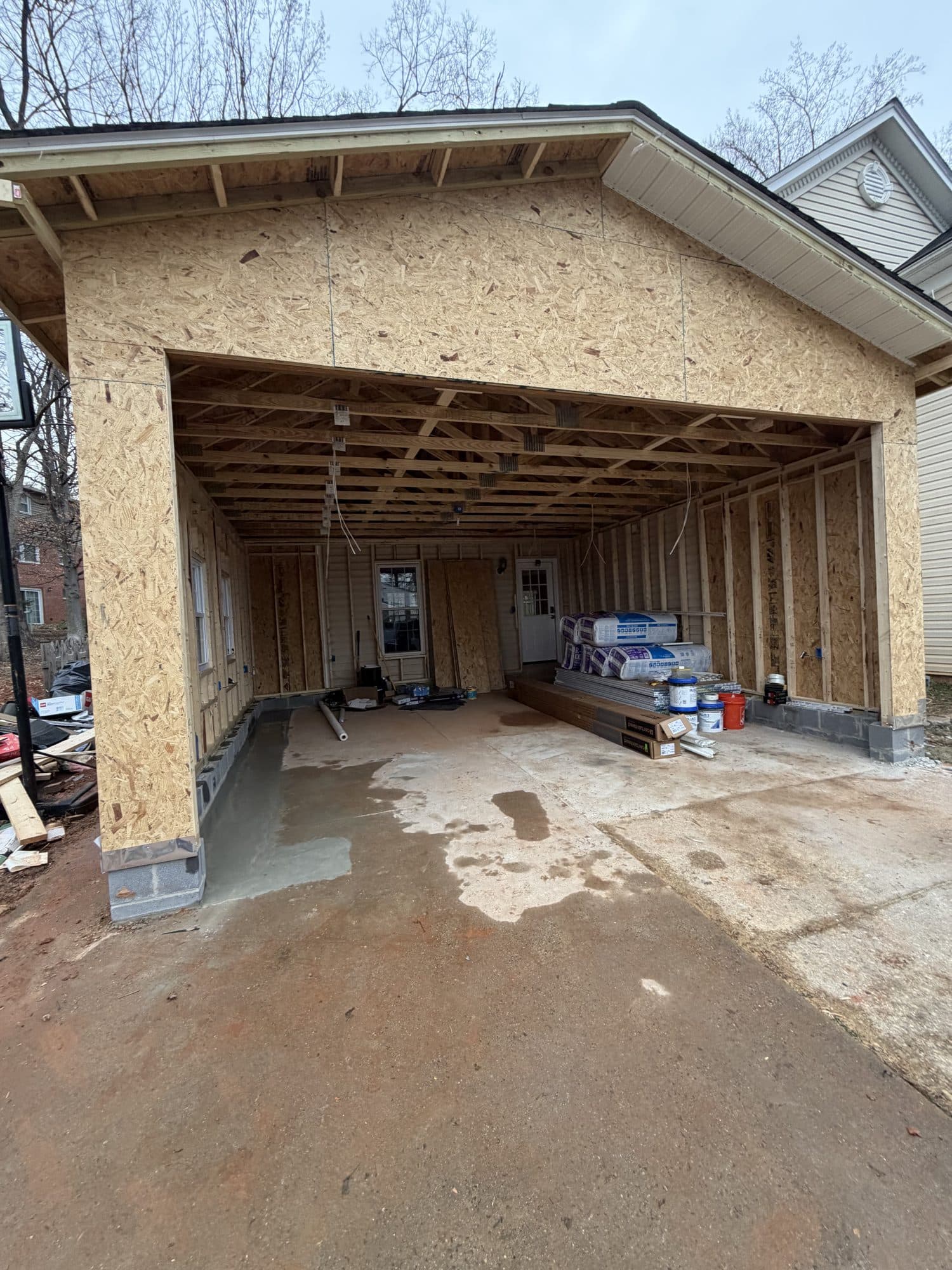 A detached garage framed in OSB sheathing with a gable and open garage bay, materials staged inside and a fresh concrete slab edge at the entrance. Garage project by Potts Brothers Construction, Arlington & Northern Virginia