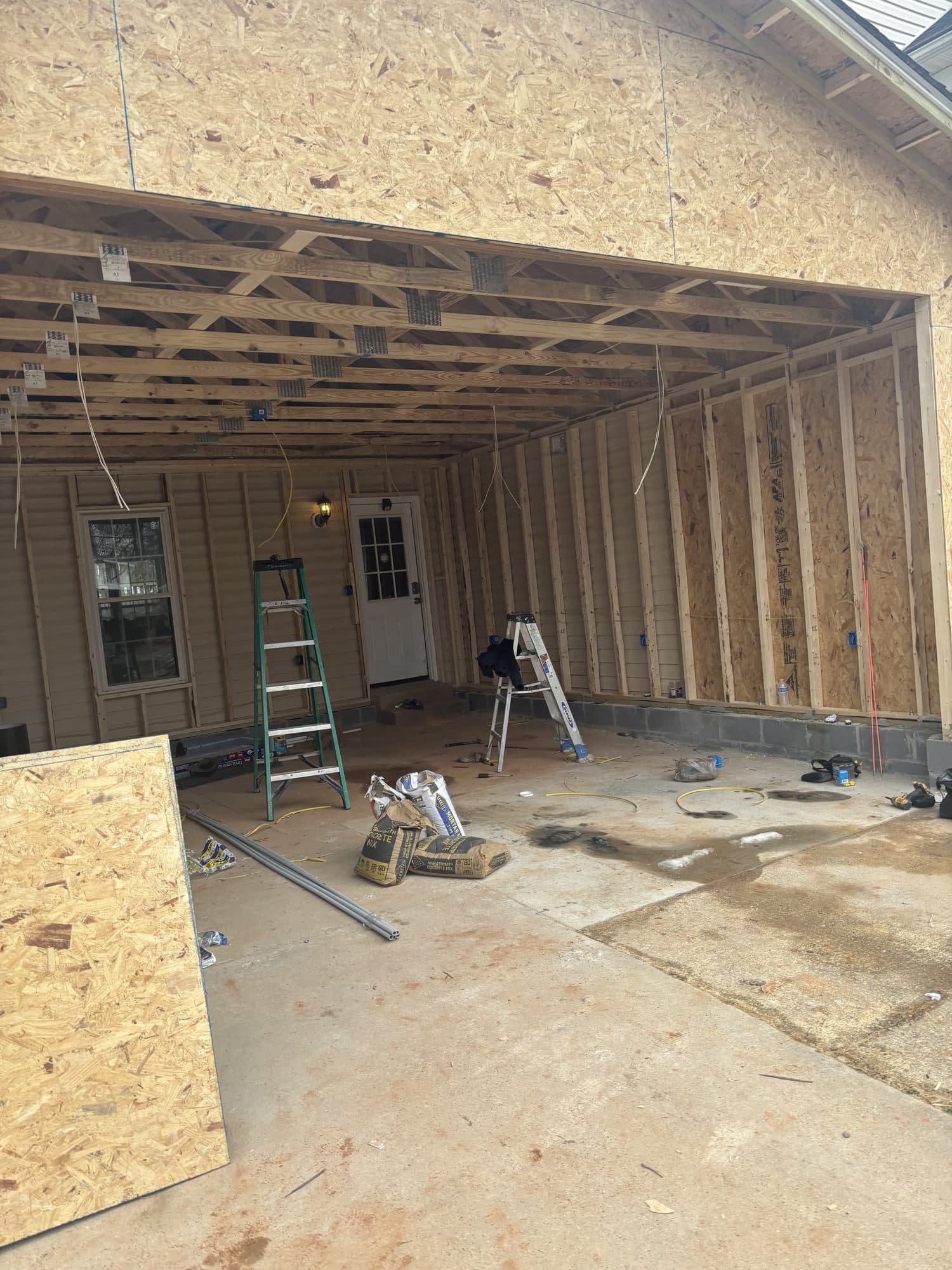 A detached garage under construction with OSB-sheathed walls and exposed rafter framing, the open garage bay revealing ladders and an interior side door. Garage project by Potts Brothers Construction, Arlington & Northern Virginia