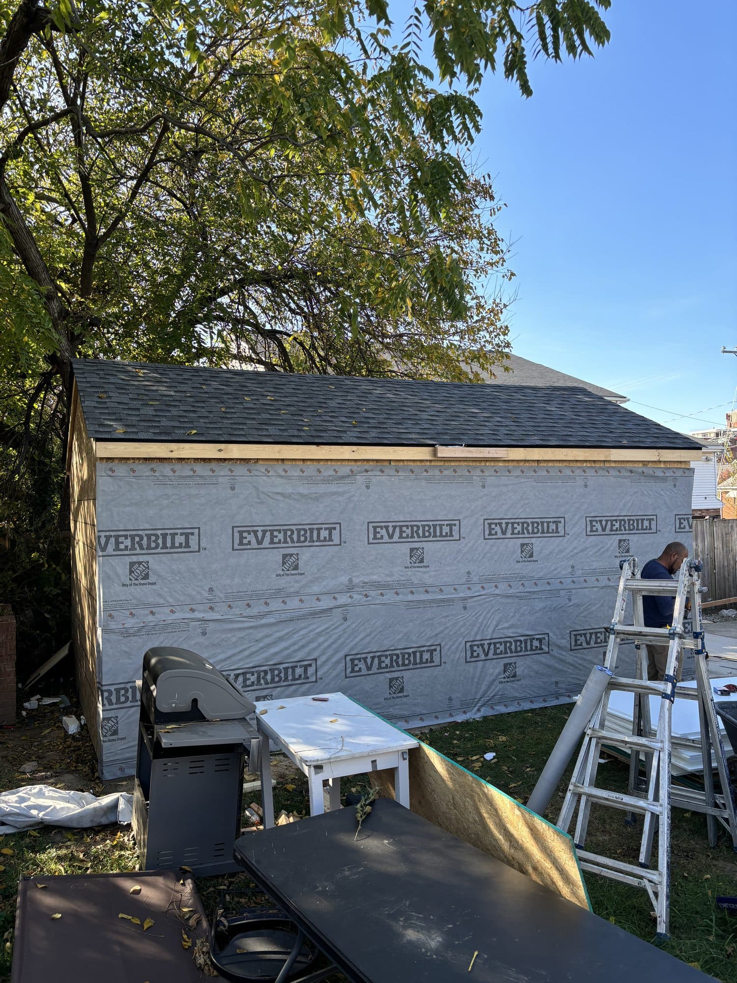 A detached garage wrapped in house wrap receives new shingles under a clear blue sky. Garage project by Potts Brothers Construction, Arlington & Northern Virginia