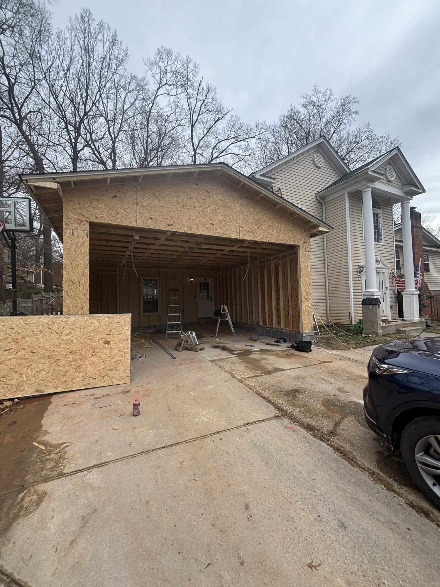 A newly framed detached garage with OSB sheathing and a gable stands beside a tan two-story home with white columns, a concrete driveway in front. Garage project by Potts Brothers Construction, Arlington & Northern Virginia