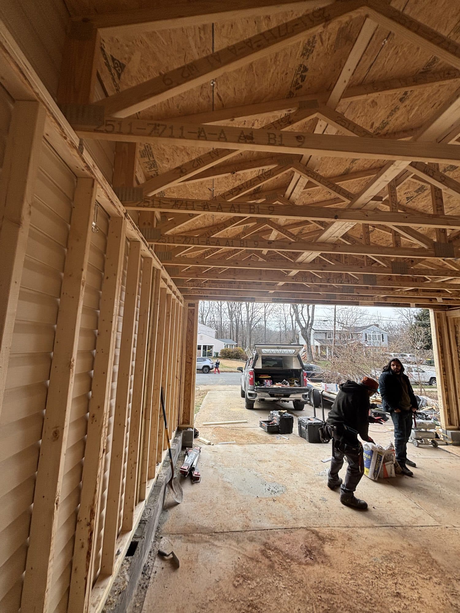 Interior of a framed garage looking out the open bay toward a driveway and parked truck, with two workers nearby. Garage project by Potts Brothers Construction, Arlington & Northern Virginia