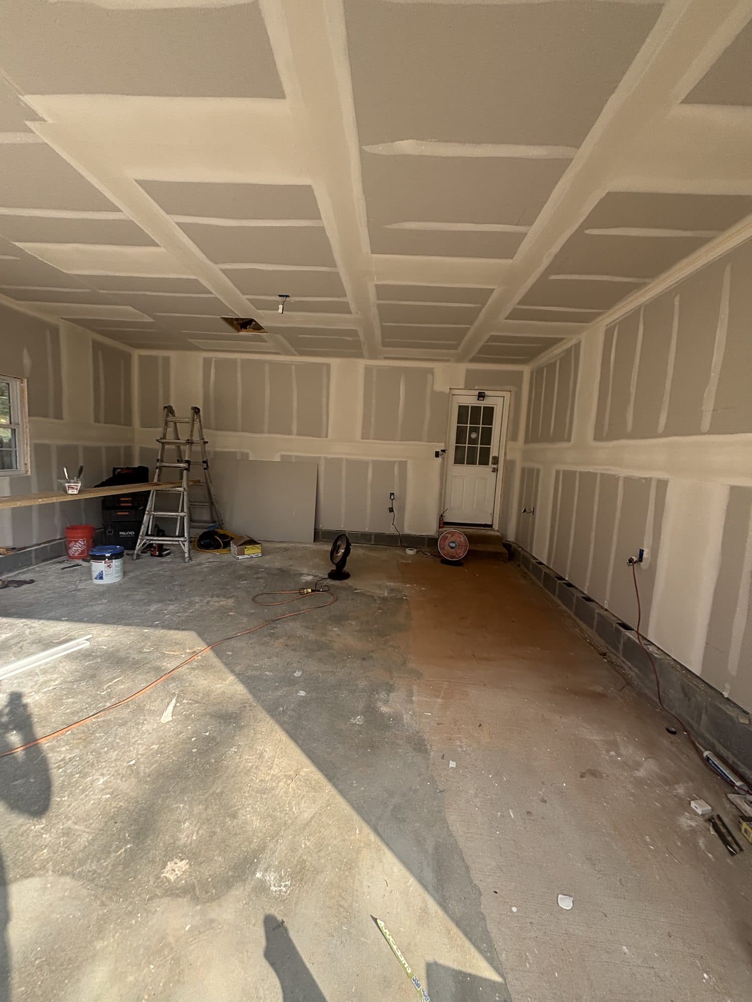 A spacious garage interior with drywall hung and seams taped on walls and ceiling, a step ladder and tools staged on the bare concrete floor. Garage project by Potts Brothers Construction, Arlington & Northern Virginia