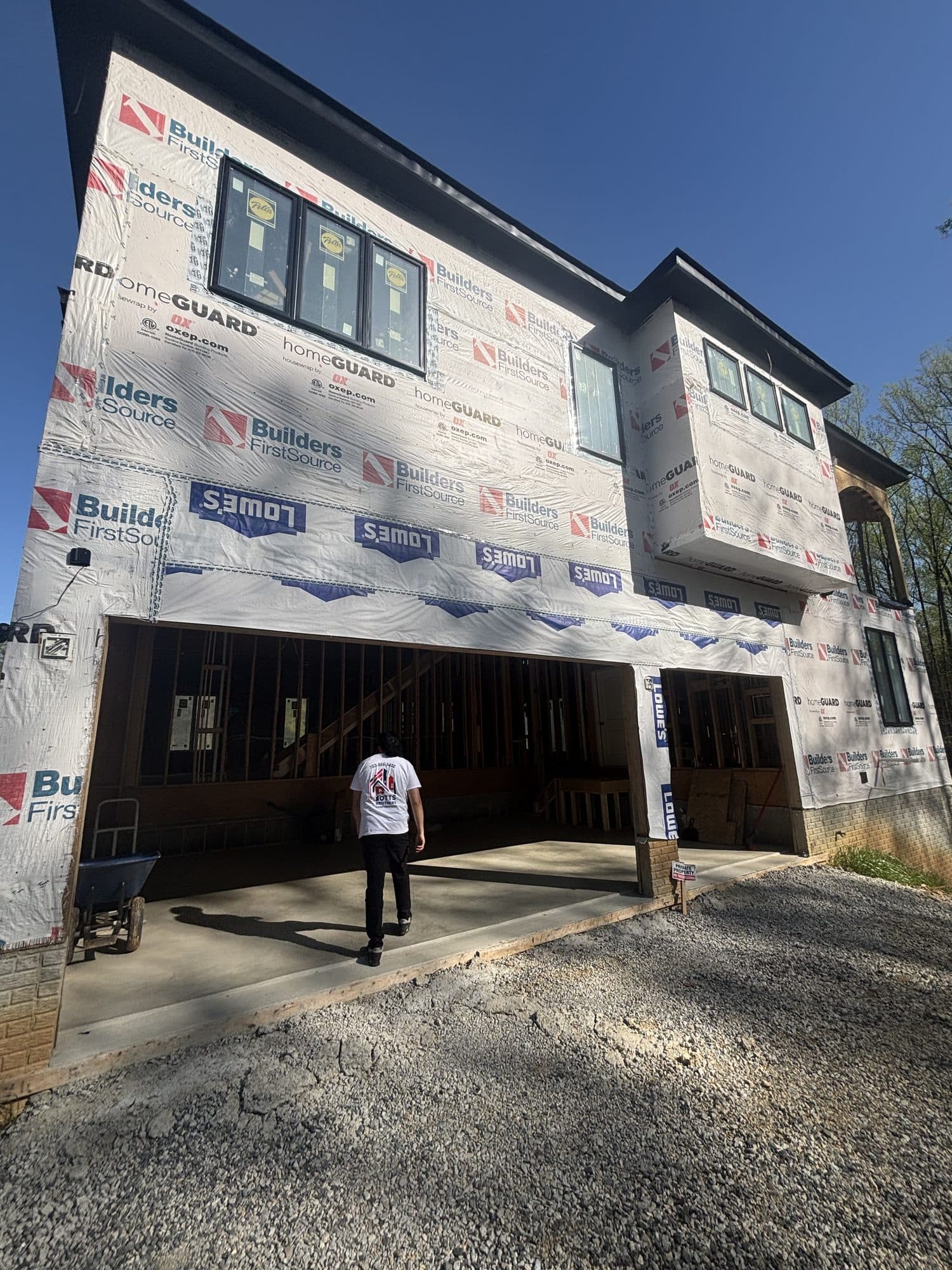 A worker approaches a modern multi-story home under construction wrapped in house wrap with double garage openings and a gravel drive. Garage project by Potts Brothers Construction, Arlington & Northern Virginia