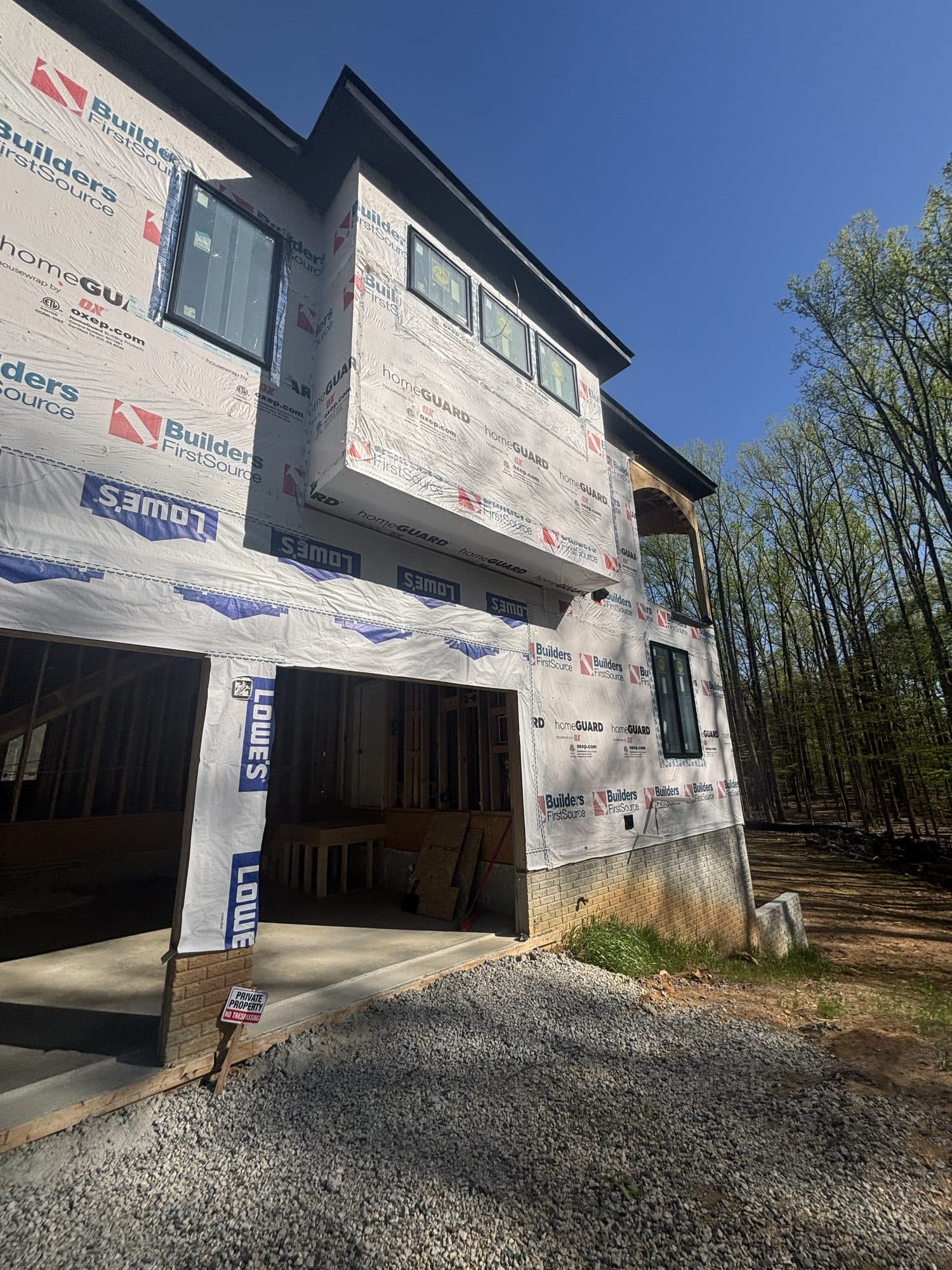 A multi-story home under construction wrapped in house wrap with a framed garage opening, fresh concrete apron, and gravel drive against a blue sky. Garage project by Potts Brothers Construction, Arlington & Northern Virginia