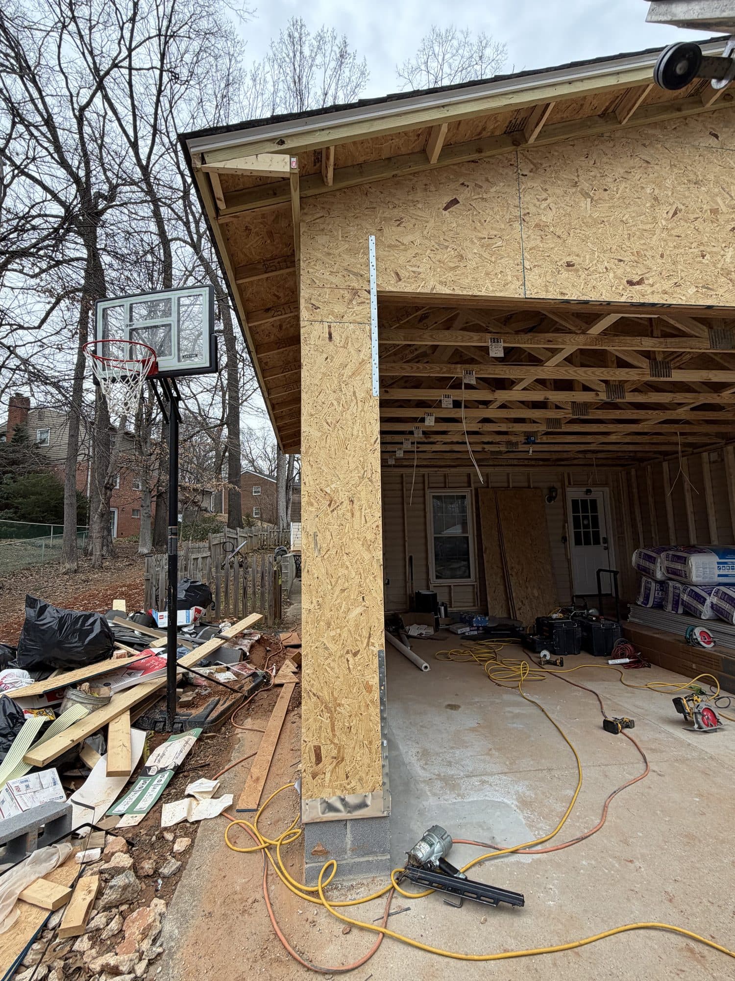 An open garage bay under construction with an OSB-sheathed corner post and exposed rafter framing beside a residential basketball hoop. Garage project by Potts Brothers Construction, Arlington & Northern Virginia