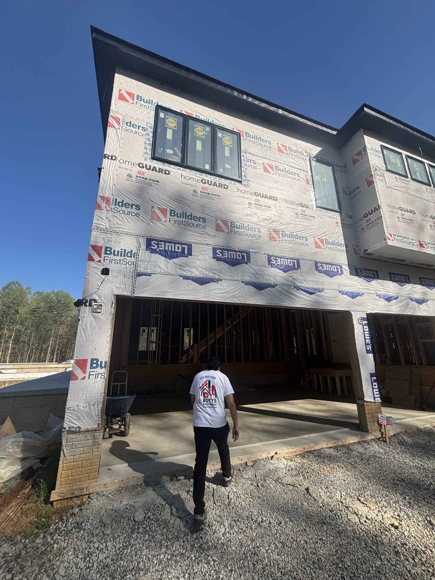A worker walks toward a three-story home under construction wrapped in house wrap with a wide garage opening and fresh concrete apron. Garage project by Potts Brothers Construction, Arlington & Northern Virginia