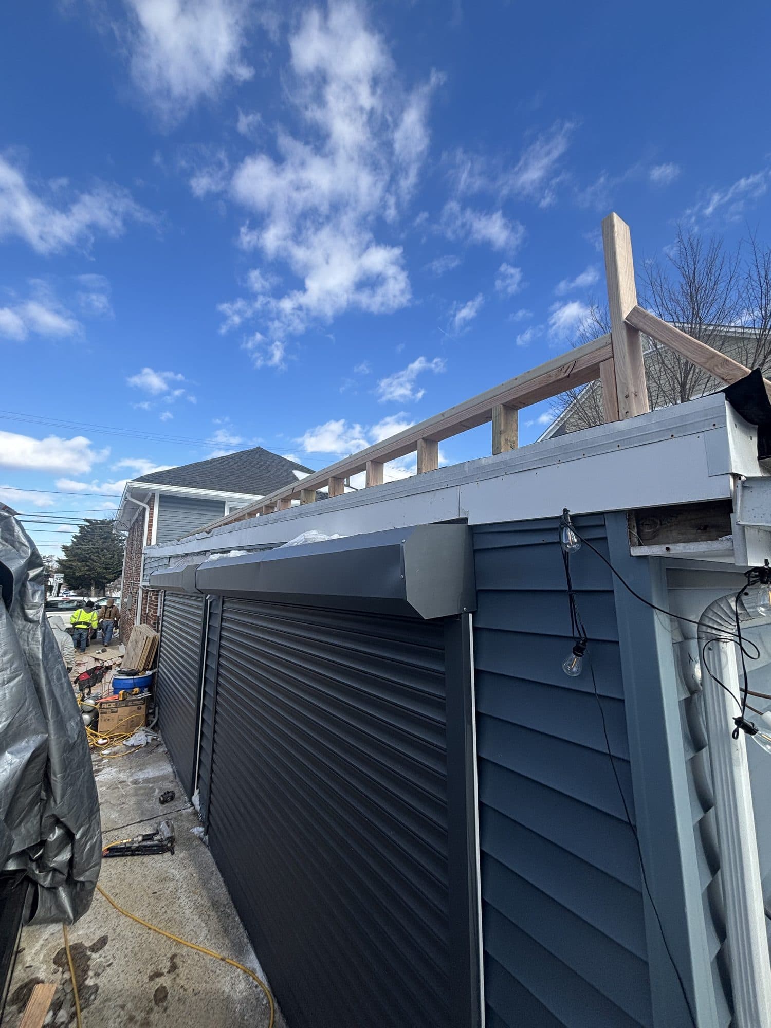A detached garage with dark roll-up doors and gray lap siding under construction, new fascia and rafter framing set against a bright blue sky. Garage project by Potts Brothers Construction, Arlington & Northern Virginia